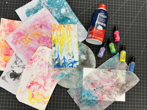 various papers with a marbling effect next to a can of shaving cream and alcohol inks