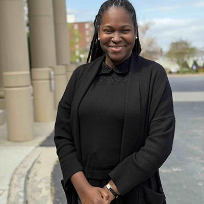 Woman wearing a black shirt and black cardigan. She is standing outdoors in a parking lot with building columns in the background.