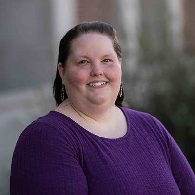 Woman in a long sleeve purple shirt with a blurred building in the background.