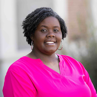 Woman in a bright pink shirt standing outdoors with a blurred brick building in the background.